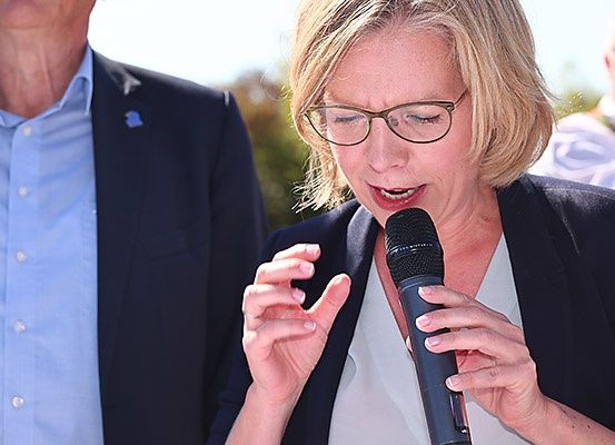 Eine Frau mit kurzen blonden Haaren und Brille spricht in ein Mikrofon. Sie gestikuliert mit ihrer rechten Hand. Hinter ihr steht ein Mann in Anzug.