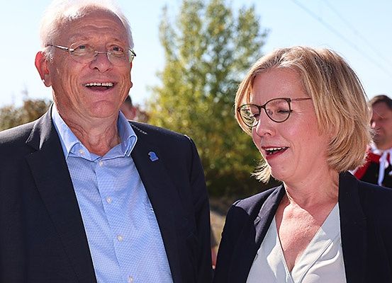 Ein älterer Mann und eine Frau stehen lächelnd zusammen, mit Bäumen und einer weiteren Person im Hintergrund.