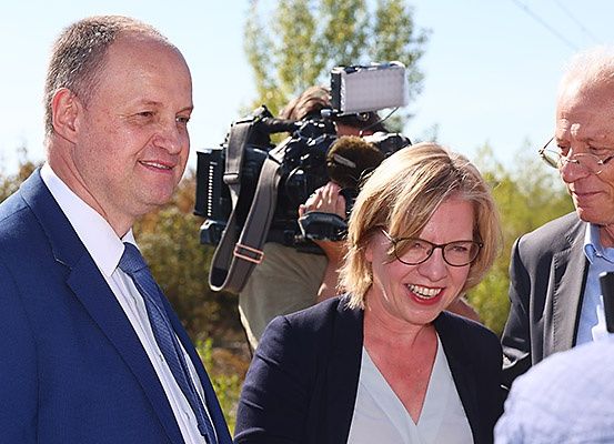 Drei Menschen lächeln auf einem Foto. Ein Mann in einem blauen Anzug und Krawatte, eine Frau in einer Brille und einem Blazer und ein älterer Mann in einer Brille. Sie werden von einer Kameracrew interviewt.