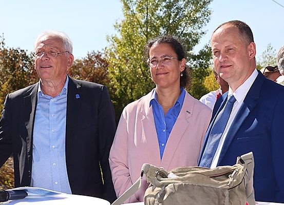 Drei Menschen stehen neben einem Tisch mit einem Mikrofon im Freien. Ein Mann auf der linken Seite trägt eine Brille und ein blaues Hemd. Eine Frau in der Mitte trägt eine Brille und einen rosa Blazer. Der Mann auf der rechten Seite trägt einen blauen Anzug.