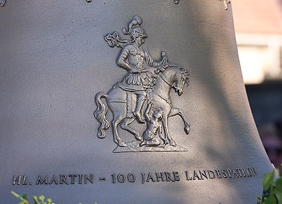 Eine Nahaufnahme einer Glocke zeigt einen Ritter auf einem Pferd, der ein Schwert hält, mit einem Kind zu seinen Füßen. Darunter steht 'HL. MARTIN ~ 100 JAHRE LANDESPATRION'.