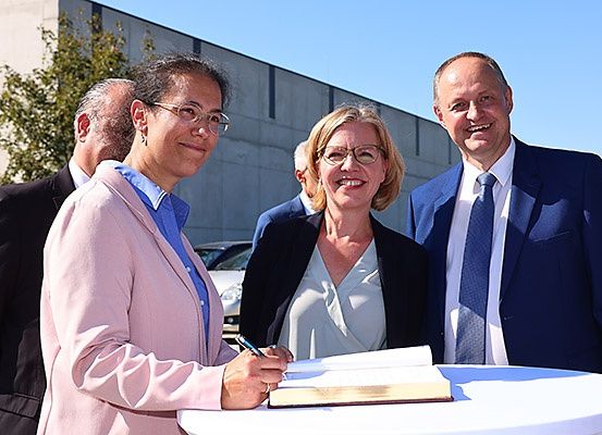 Eine Gruppe von Menschen steht vor einem Gebäude, wobei zwei Frauen ein Dokument auf einem Tisch unterschreiben, während andere zusehen.