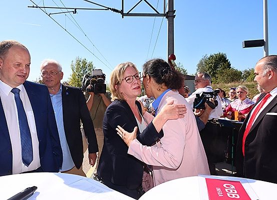 Eine Gruppe von Menschen, darunter ein Mann und eine Frau mit Brille, steht draußen mit einem Tisch und einem Banner mit der Aufschrift 'GBB'. Sie sind von Journalisten mit Kameras umgeben.