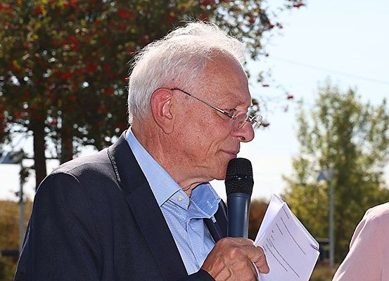 Ein älterer Mann mit weißen Haaren und einer Brille spricht in ein Mikrofon, während er ein Blatt Papier in der Hand hält.