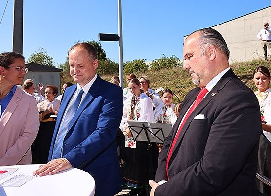 Eine Gruppe von Menschen steht um einen Tisch herum, wobei zwei Männer in Anzügen sprechen. Dahinter spielen Menschen in traditioneller Kleidung Instrumente. Eine Frau mit Brille steht links.