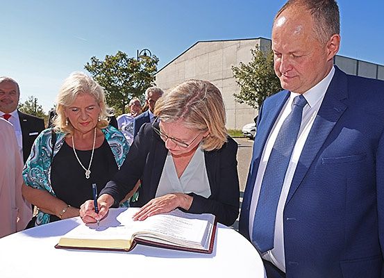 Drei Personen, ein Mann und zwei Frauen, unterzeichnen ein Dokument auf einem Tisch. Die unterzeichnende Frau trägt eine Brille. Hinter ihnen stehen mehrere Personen, einige von ihnen tragen Anzüge. Das Gebiet ist von Bäumen umgeben.