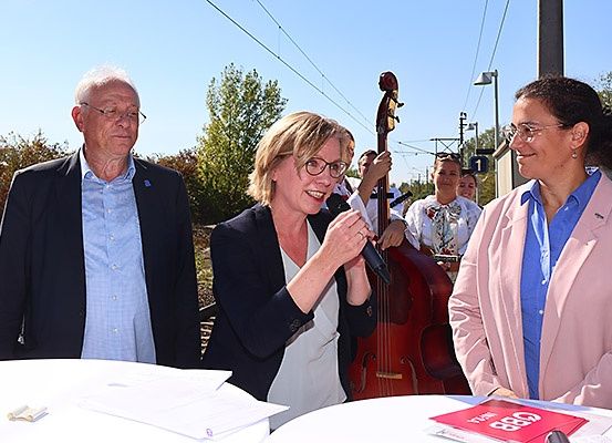 Drei Personen stehen auf einer Plattform mit einem Tisch, wahrscheinlich bei einer Veranstaltung. Eine Frau hält ein Mikrofon und lächelt, während ein Mann hinter ihr steht. Dahinter spielt ein Cellist, und andere sind in der Nähe. Bäume und eine Bahnlinie sind im Hintergrund.