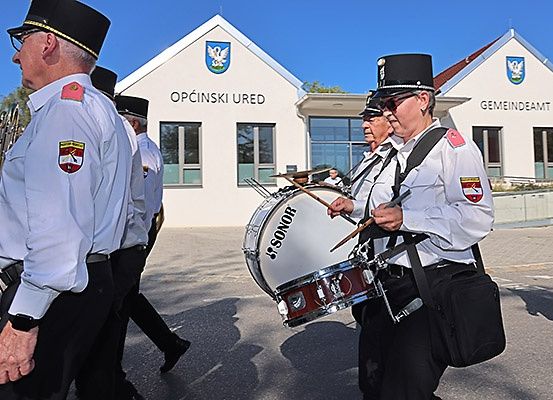 Eine Gruppe uniformierter Männer spielt Musikinstrumente vor einem weißen Gebäude mit der Aufschrift Opcinski Ured und einem Logo.