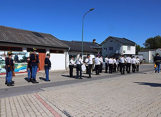 Eine Gruppe uniformierter Personen steht auf einer Straße mit Ziegelbelag und hält Gewehre und Trommeln. Sie scheinen eine Marschkapelle zu sein.