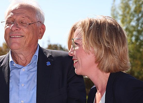 Ein älterer Mann und eine junge Frau stehen nah beieinander, beide lächeln und schauen sich an. Der Mann trägt eine Brille und einen Anzug. Die Frau hat kurze blonde Haare und eine Brille. Sie stehen draußen mit Bäumen im Hintergrund.