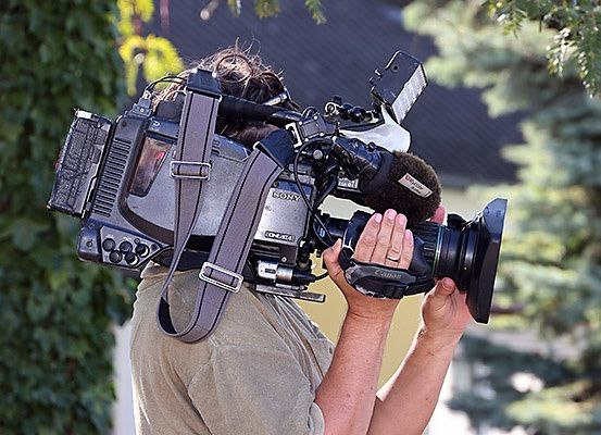 Eine Person in einem braunen Hemd hält eine Sony-Videokamera mit einem Mikrofon vor sich.