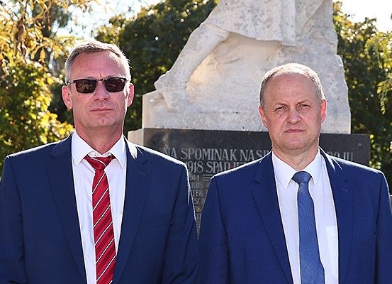 Zwei Männer stehen Seite an Seite vor einem großen Steinmonument mit Inschriften, gekleidet in formelle Anzüge und Krawatten, mit Bäumen im Hintergrund.