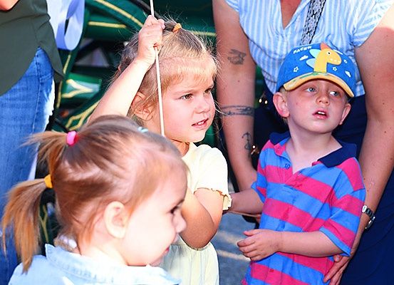 Drei Kinder, eines mit einem Stock, schauen etwas an, wobei eine Frau die Hand des Jungen hält.
