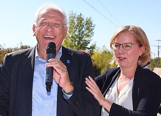 Ein älterer Mann hält ein Mikrofon und lächelt, während eine Frau mit Brille neben ihm steht und ebenfalls lächelt. Sie sind beide formell gekleidet. Hinter ihnen befinden sich Bäume und eine Stromleitung im Hintergrund.