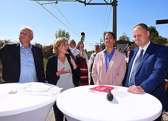 Mehrere Personen stehen um Tische herum bei einer Veranstaltung im Freien. Eine Frau gestikuliert, während sie spricht. Ein Mikrofon und Papiere liegen auf einem Tisch. Ein Mann hält ein Cello. Elektrische Leitungen sind im Hintergrund.