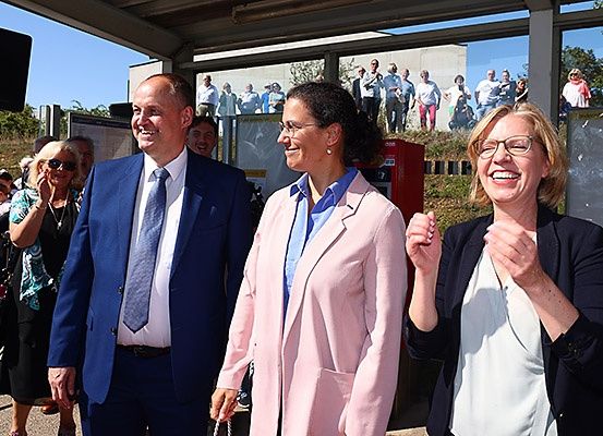 Drei Personen, zwei Frauen und ein Mann, lächeln und klatschen bei einer Veranstaltung im Freien, mit einer Menge im Hintergrund.