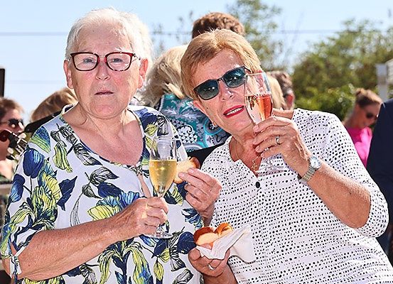 Zwei Frauen, beide mit Brille, halten Weinglas und Essen und lächeln für ein Foto. Dahinter stehen mehrere Menschen und schauen in verschiedene Richtungen.