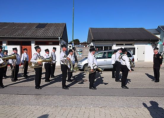 Eine Brassband spielt auf einer Straße, mehrere Personen stehen vor Gebäuden und ein silbernes Auto ist geparkt.