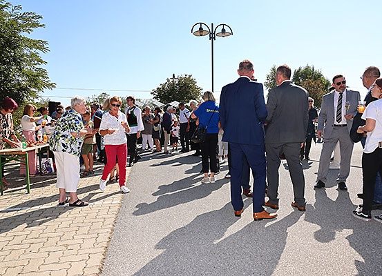 Eine Menschenmenge versammelt, stehend und im Gespräch. Einige tragen Sonnenbrillen und halten Getränke. Eine Frau serviert Getränke von einem Tisch. Straßenlaternen und Bäume sind im Hintergrund.