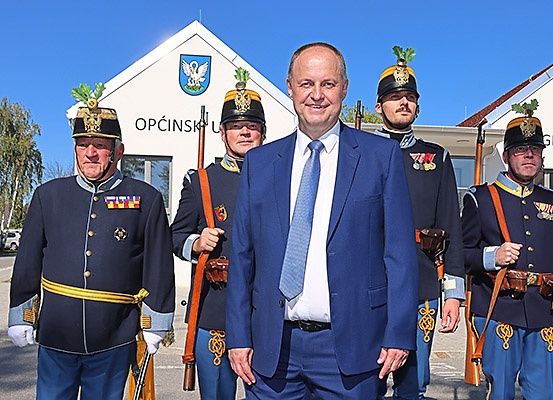 Ein Mann im Anzug steht mit drei Militärangehörigen, zwei mit Gewehr und einer mit einem Schwert, vor einem Gebäude mit einem Schild, auf dem Opcinski steht.