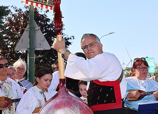 Ein Mann in traditioneller Kleidung spielt eine große Geige, umgeben von einer Gruppe von Musikern. Sie spielen im Freien an einem sonnigen Tag.