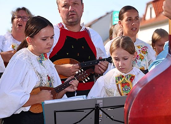 Eine Gruppe von Menschen singt und spielt Instrumente, darunter eine Frau, die eine Gitarre und ein Mann, der eine Mandoline spielt, mit anderen in traditioneller Kleidung.