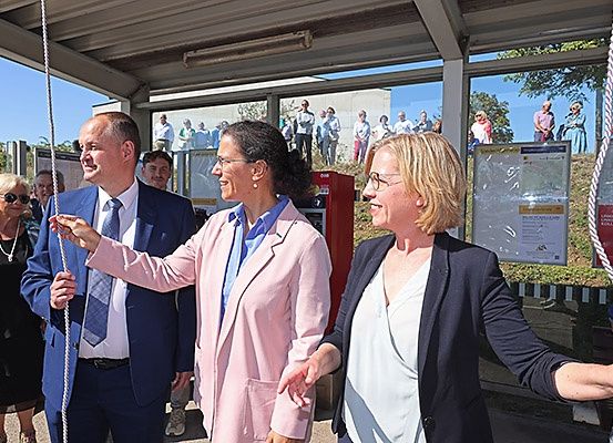 Drei Erwachsene, zwei Frauen und ein Mann, stehen in einem Schutzraum und lächeln. Im Hintergrund stehen mehrere Personen auf einem Hügel.