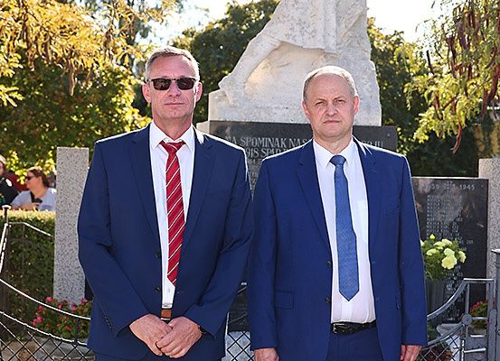 Zwei Männer in Anzügen stehen vor einem Denkmal mit einer Statue, möglicherweise auf einem Friedhof. Hinter ihnen stehen einige Menschen und sehen sich etwas an.