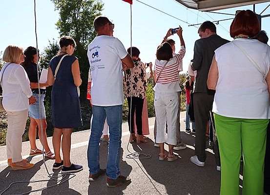 Eine Gruppe von Menschen steht am Straßenrand und macht Fotos mit ihren Handys. Ein Mann in einem weißen T-Shirt mit Logo schaut nach links.