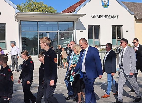 Mehrere Personen in formeller Kleidung gehen vor einem Gebäude mit dem Schild 'Gemeindeamt'. Unter ihnen sind einige Kinder in Uniformen. Hinter ihnen unterhält sich ein Mann mit einer Frau. Im Hintergrund stehen Personen und sehen sich etwas an.