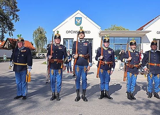 Fünf Männer in Uniform, mit Gewehren in der Hand, stehen vor einem Gebäude mit der Aufschrift 'OPICINSRED'. Sie tragen Hüte mit goldenen Federn und Gürtel. Hinter ihnen steht eine Person in einem grünen Kleid. Im Hintergrund befinden sich ein Baum und ein Haus.