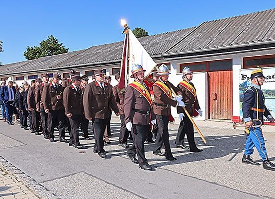 Eine Blaskapelle in Uniformen zieht die Straße entlang, gefolgt von Zuschauern. Ein Trompeter führt die Kapelle an.