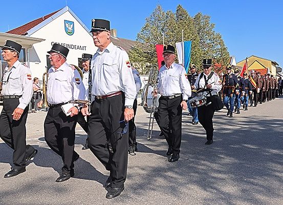Eine Gruppe von Männern in Uniform marschiert auf der Straße, einige mit Musikinstrumenten. Ihnen folgt eine Reihe von Soldaten. Gebäude und Bäume säumen die Straße.