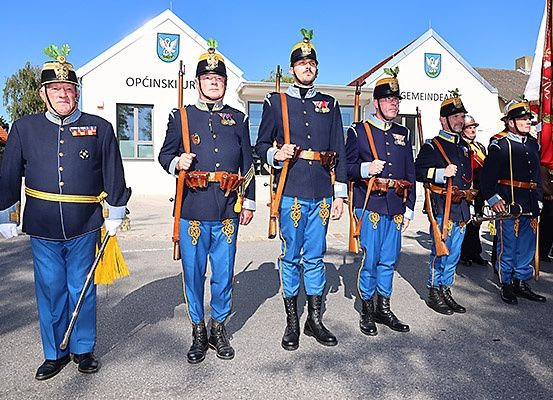 Sechs uniformierte Männer stehen in einer Reihe, jeder mit einem Gewehr in der Hand. Sie stehen vor einem Gebäude mit weißer Fassade und einem Wappen.