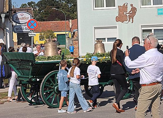 Eine Gruppe von Menschen, die um einen geschmückten Wagen mit Glocken herumgeht, auf einem Stadtplatz, mit Gebäuden und Grünpflanzen im Hintergrund.