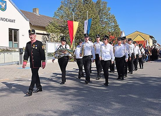Eine Musikkapelle, angeführt von einem älteren Mann, zieht in Uniform spielend Musikinstrumente spielend die Straße entlang. Dahinter stehen zwei Fahnenmasten mit Fahnen. Gebäude säumen die Straße.
