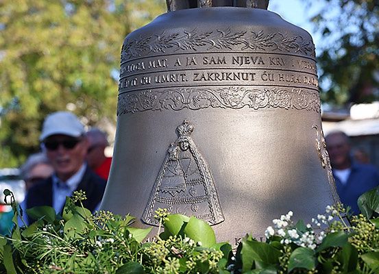 Eine große Glocke, verziert mit Blättern und einer Frauenfigur, ist mit Grünzeug geschmückt und wird von Menschen in einem unscharfen Hintergrund besucht.