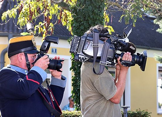Zwei Männer vor einem Gebäude mit einem Baum, einer in Uniform und mit einer Kamera, der andere in einem grauen Shirt und mit einer großen Videokamera.