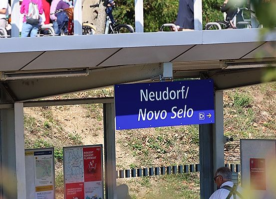 Ein blaues Schild mit der Aufschrift 'Neudorf/Novo Selo' hängt an einer Plattform über einem Bahnhof. Menschen stehen auf der Plattform, und Fahrräder sind geparkt. Ein Mann geht vor dem Schild.