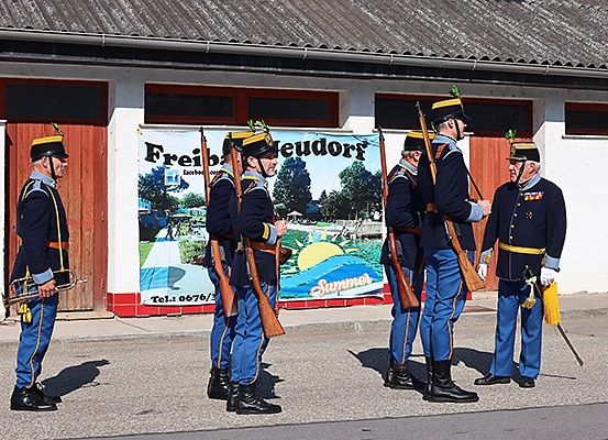Fünf Männer in Uniform stehen auf der Straße, Gewehre in der Hand, vor einem Gebäude mit einem Banner, das Freib Neudorf liest.
