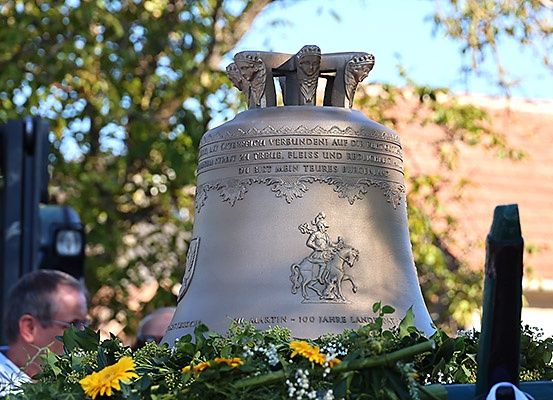 Eine Glocke mit einem Blumenkranz steht auf einem Wagen. Ein Mann mit Brille steht im Hintergrund. Die Glocke ist mit einem Reiter auf einem Pferd und etwas Text graviert. Der Himmel ist klar.
