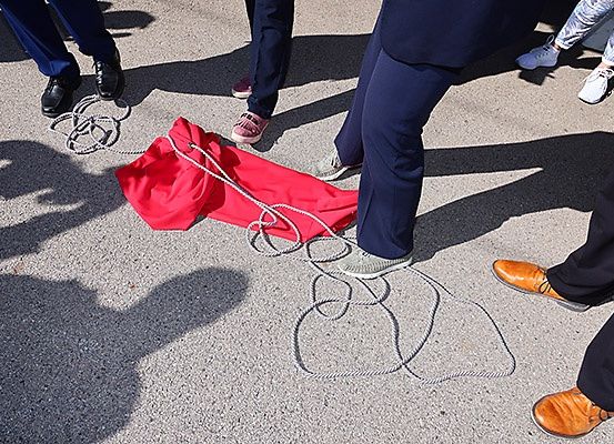 Fünf Personen stehen auf einer Betonfläche; ein roter Stoff liegt auf dem Boden, mit Seilen gebunden. Sie tragen verschiedene Schuhe, und Schatten fallen auf die Oberfläche.