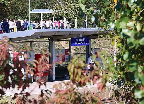 Menschen versammeln sich auf einer überdachten Plattform des Bahnhofs Neudorf/Novo Selo, umgeben von Bäumen und Laub.