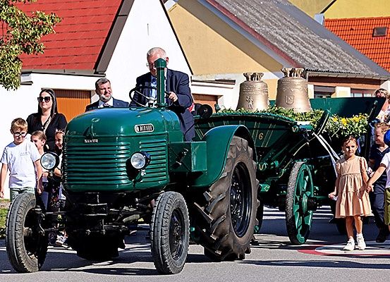 Ein grüner Oldtimer-Traktor mit Glocken wird von einem Mann in Anzug gefahren, während Kinder und Erwachsene an der Straßenseite zusehen.