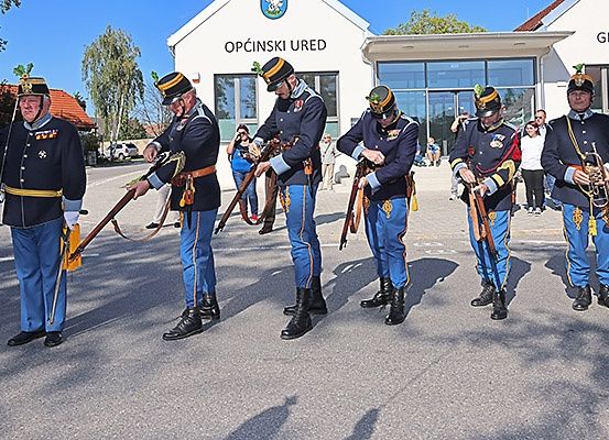 Fünf Männer in Militäruniformen führen eine Zeremonie vor einem Gebäude durch. Sie halten Gewehre und tragen Hüte. Dahinter stehen Menschen, die Fotos machen.