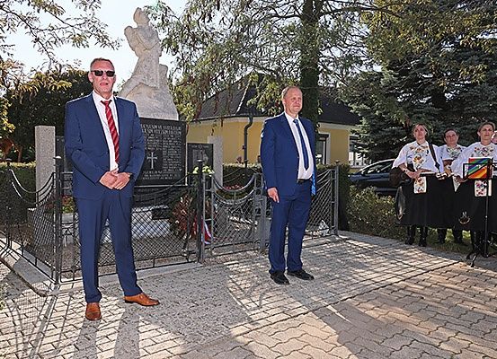 Zwei Männer in Anzügen stehen vor einem Denkmal mit einer Statue, flankiert von Frauen in traditioneller Kleidung.