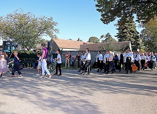 Eine Parade mit Kindern und Erwachsenen, einige mit Gitarren, geht auf einem Schotterweg unter klarem Himmel.