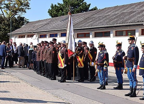 Eine Gruppe von Militäroffizieren steht in Formation mit einer Flagge, während sich Zuschauer dahinter versammeln. Sie tragen Hüte und Uniformen, und das Gebäude dahinter hat ein graues Dach.