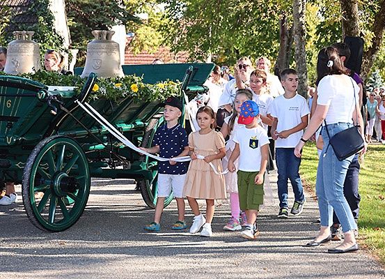 Mehrere Kinder und Erwachsene halten ein Seil vor einem dekorierten grünen Wagen mit Glocken oben. Sie scheinen an einer Parade teilzunehmen.