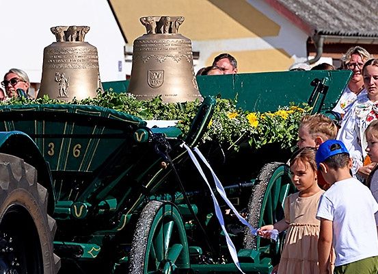 Ein geschmückter Traktor mit Glocken und Blumen wird von einer Gruppe von Menschen gezogen. Einige tragen Hüte und Brillen. Sie befinden sich in einem Außenbereich.